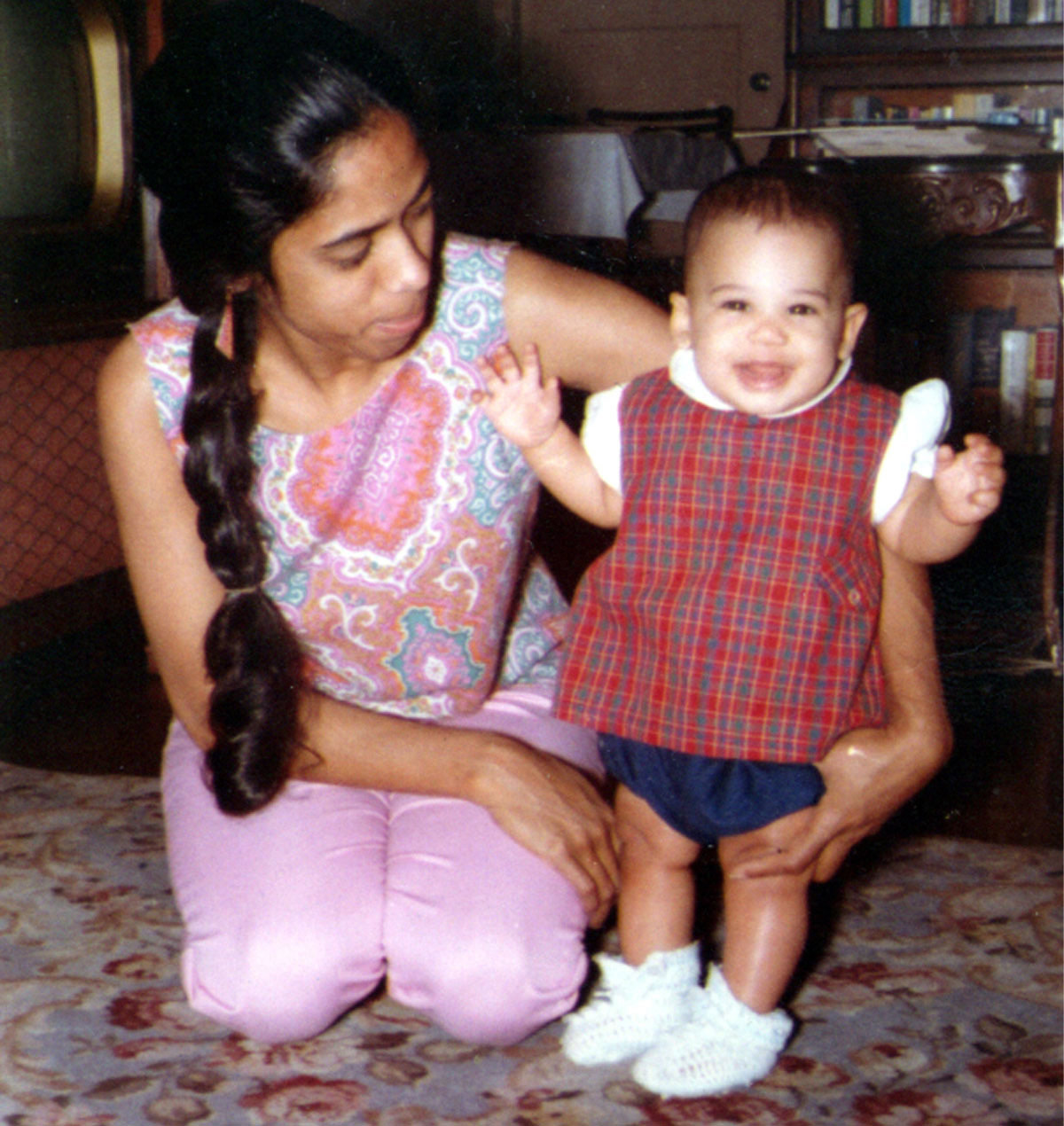 Kamala cuando era una niña pequeña vistiendo un suéter a cuadros rojo y zapatos de punto blanco, sonriendo junto a su madre, Shyamala.